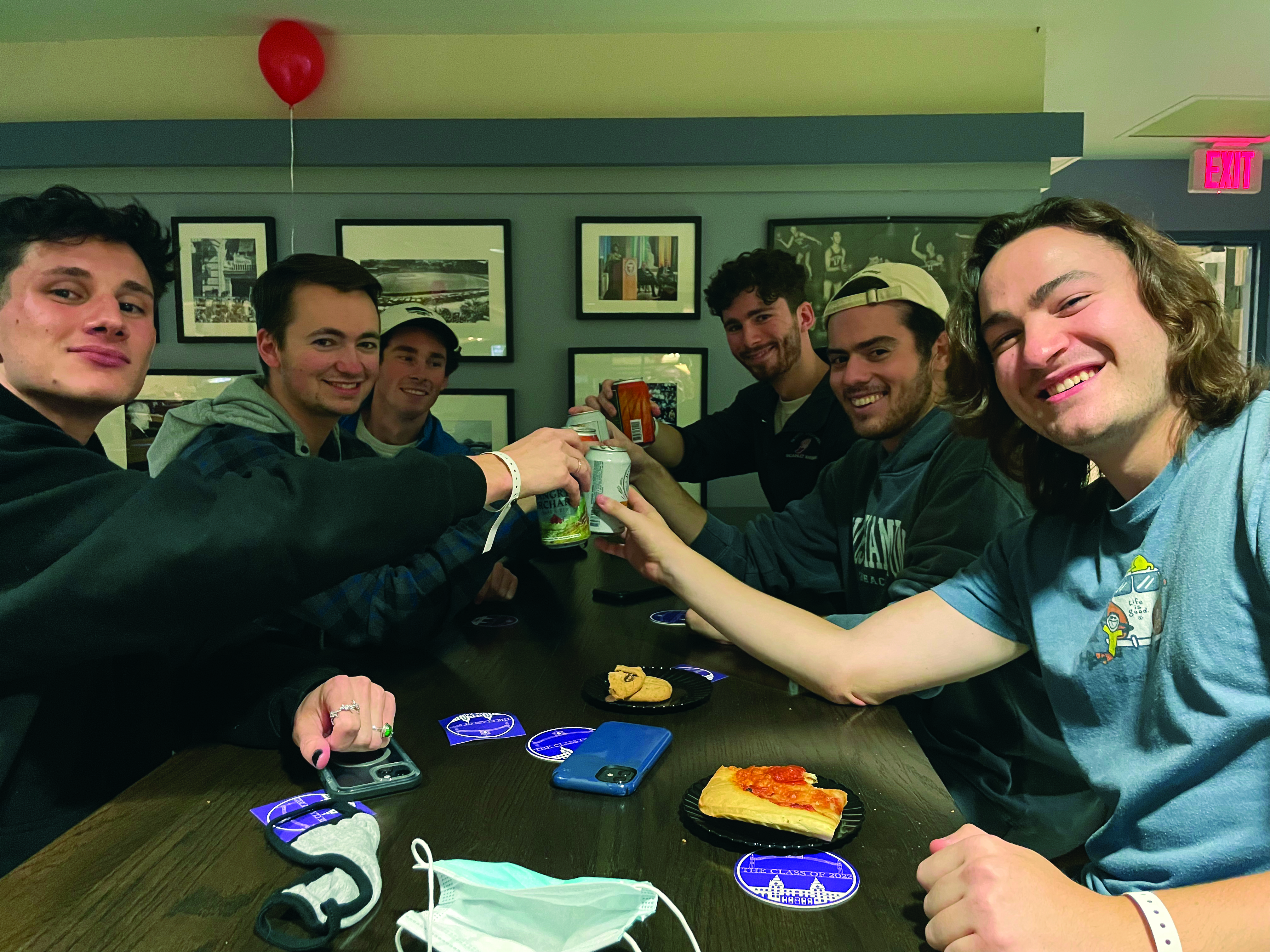 A group of six young men stand around a table and hold their drinks together for a toast