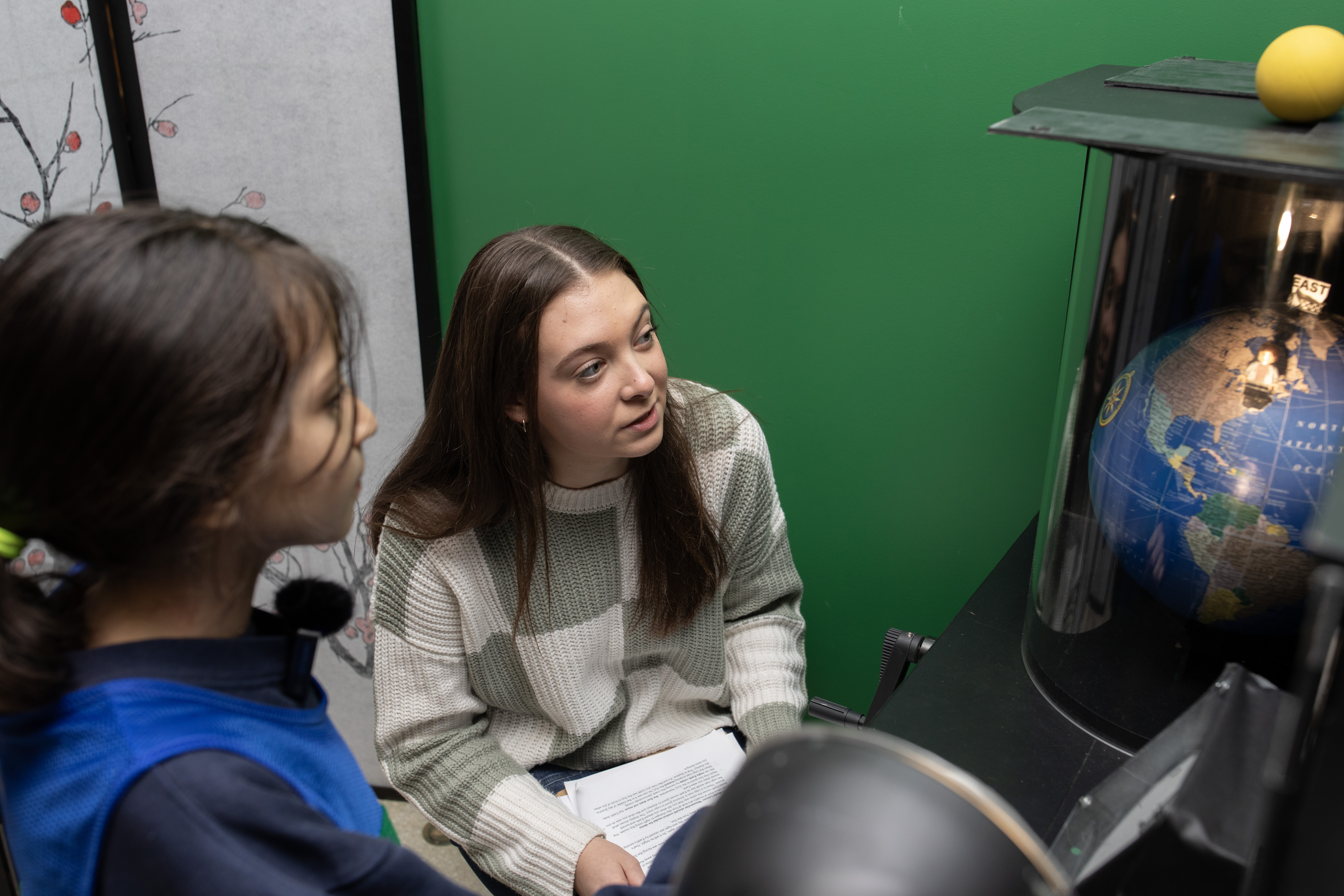 A female college students crouches down next to an elementary school girl to look at a globe.