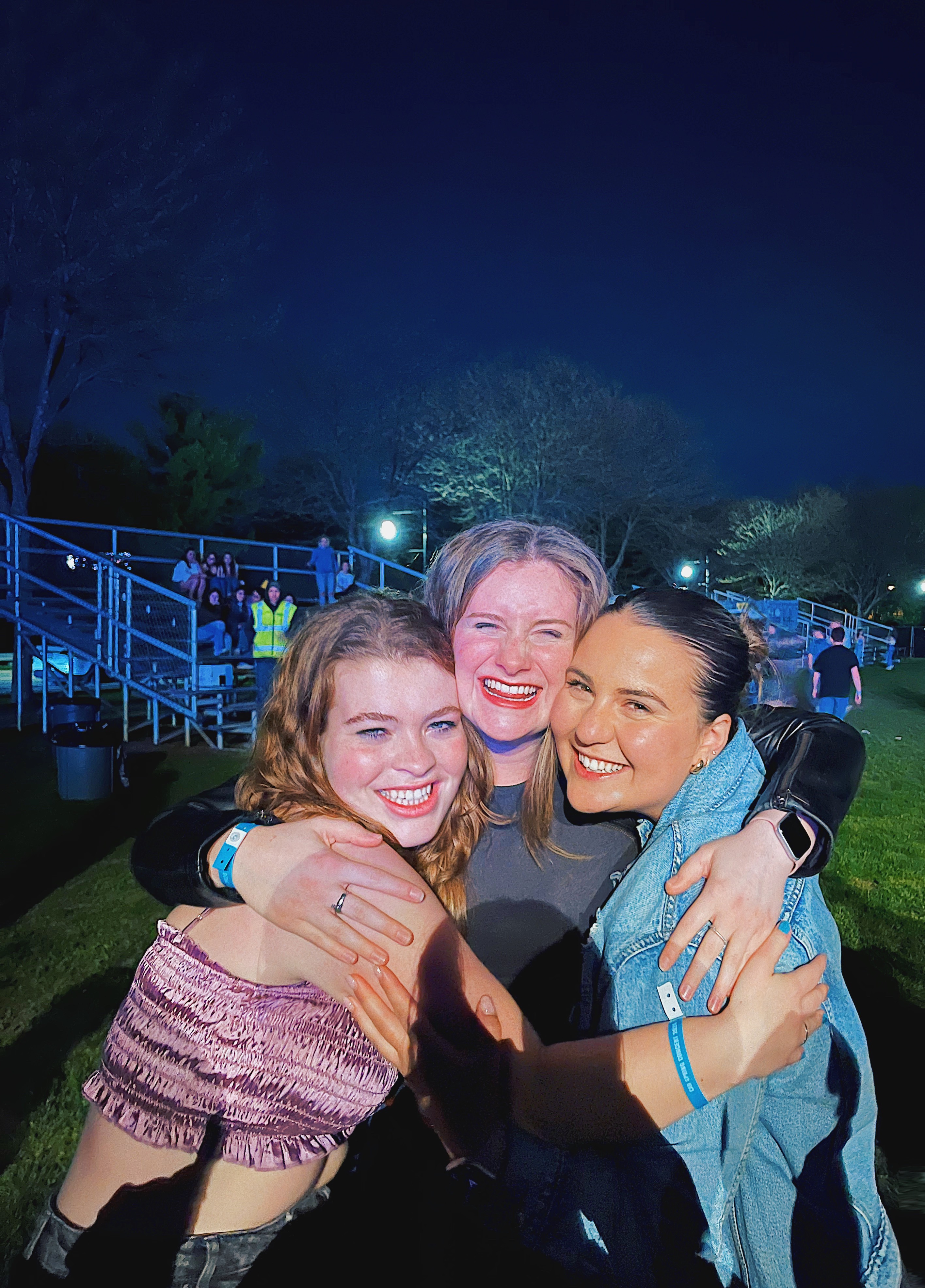 Three young women pose huddled together and smiling at night with bleachers in the background