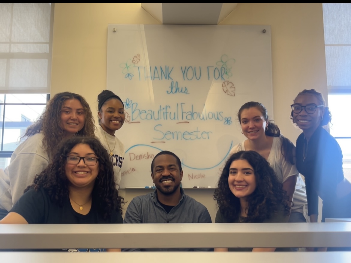 A group of students gather around their teacher smiling with a white board behind them reading &quot;Thank you for this beautiful semester&quot;
