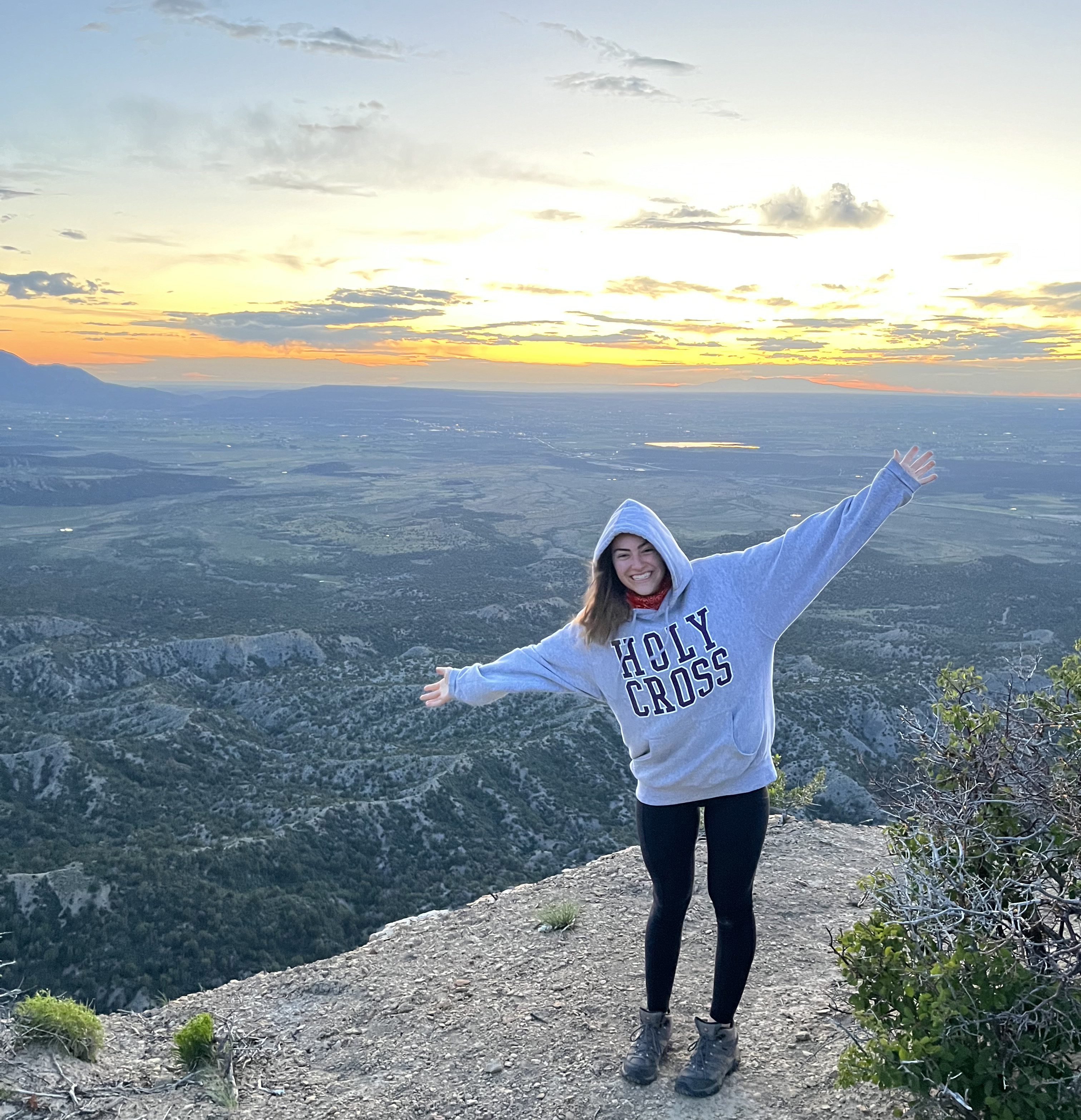 A young woman wearing a hooded Holy Cross sweatshirt stands atop a mountain overlooking a view of the valley