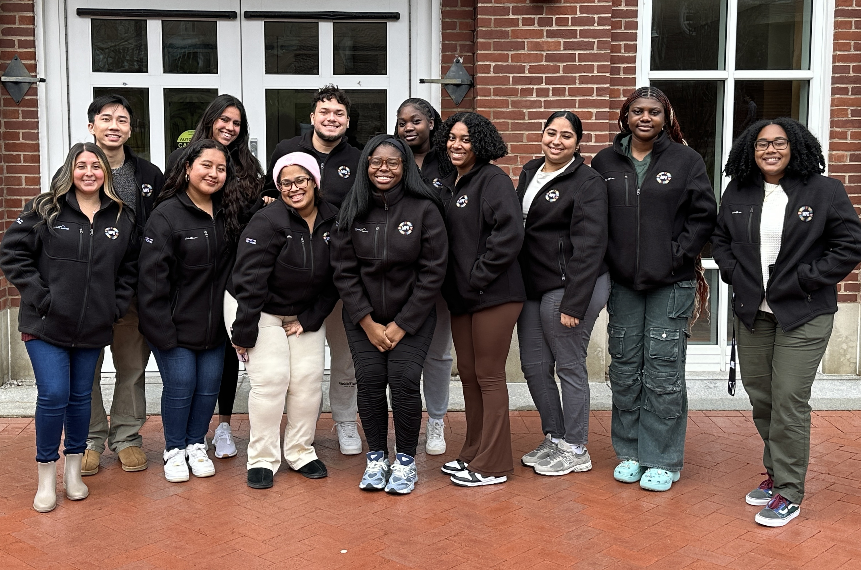 A group of students huddle together smiling outside a brick building