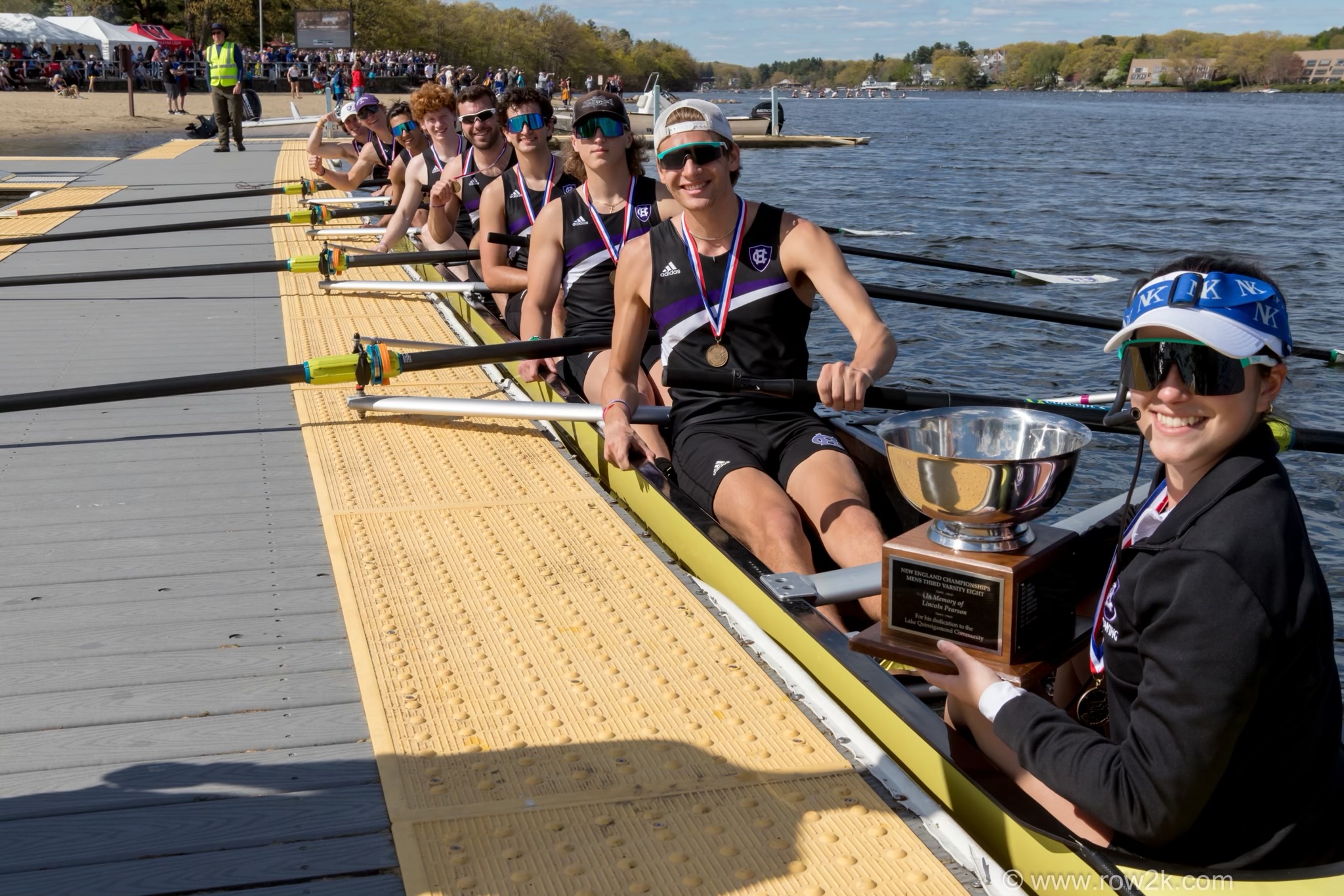 A Holy Cross rowing team sits in the boat along the dock while the first person holds a trophy