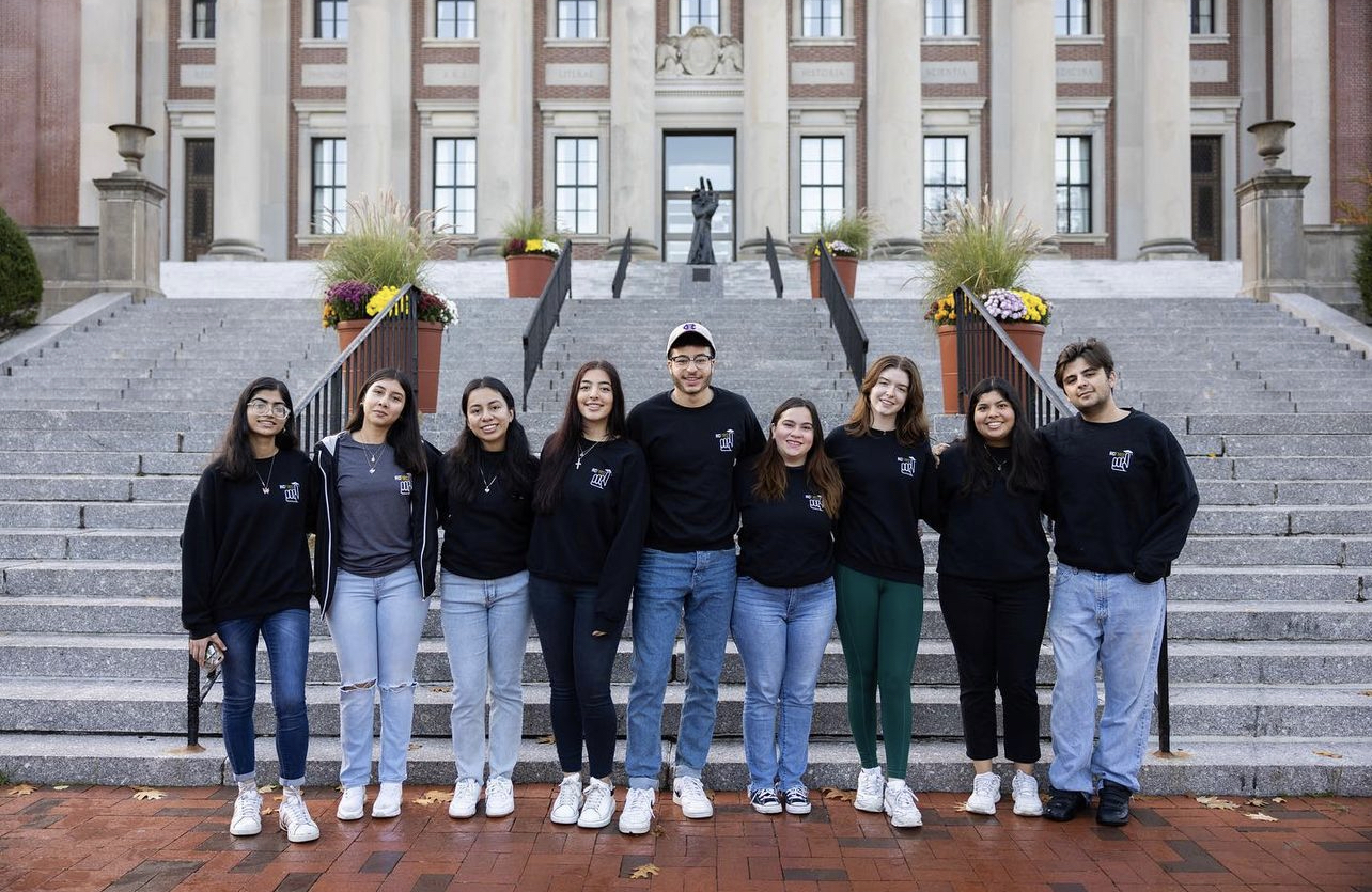 A group of students, the executive board of HCF1RST, stands at the steps of Dinand Library