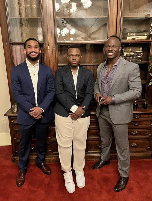 Three young men in formal attire pose with a bookshelf behind them