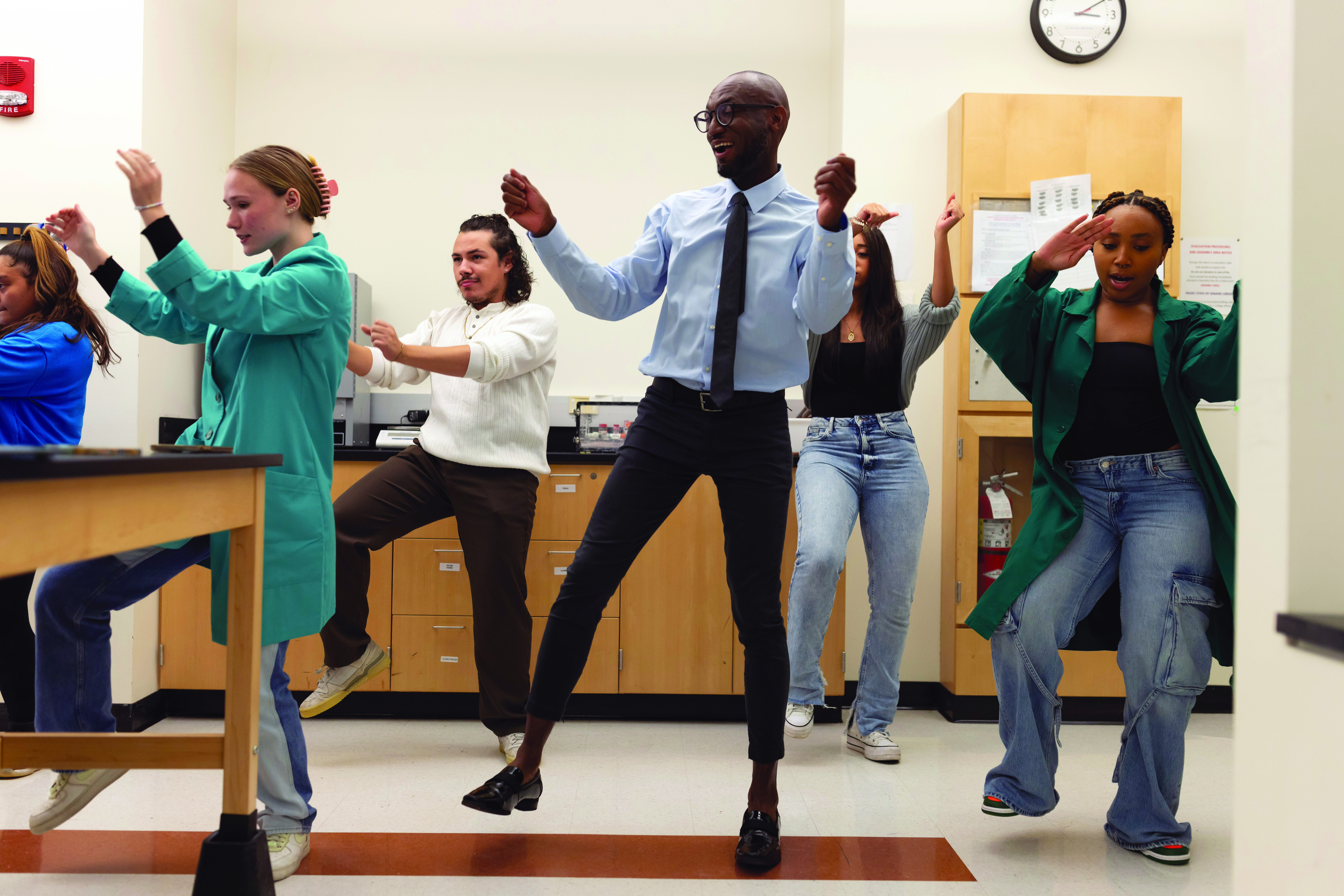 A man and four college students next to and behind him dance with their arms raised and right foot in the air.