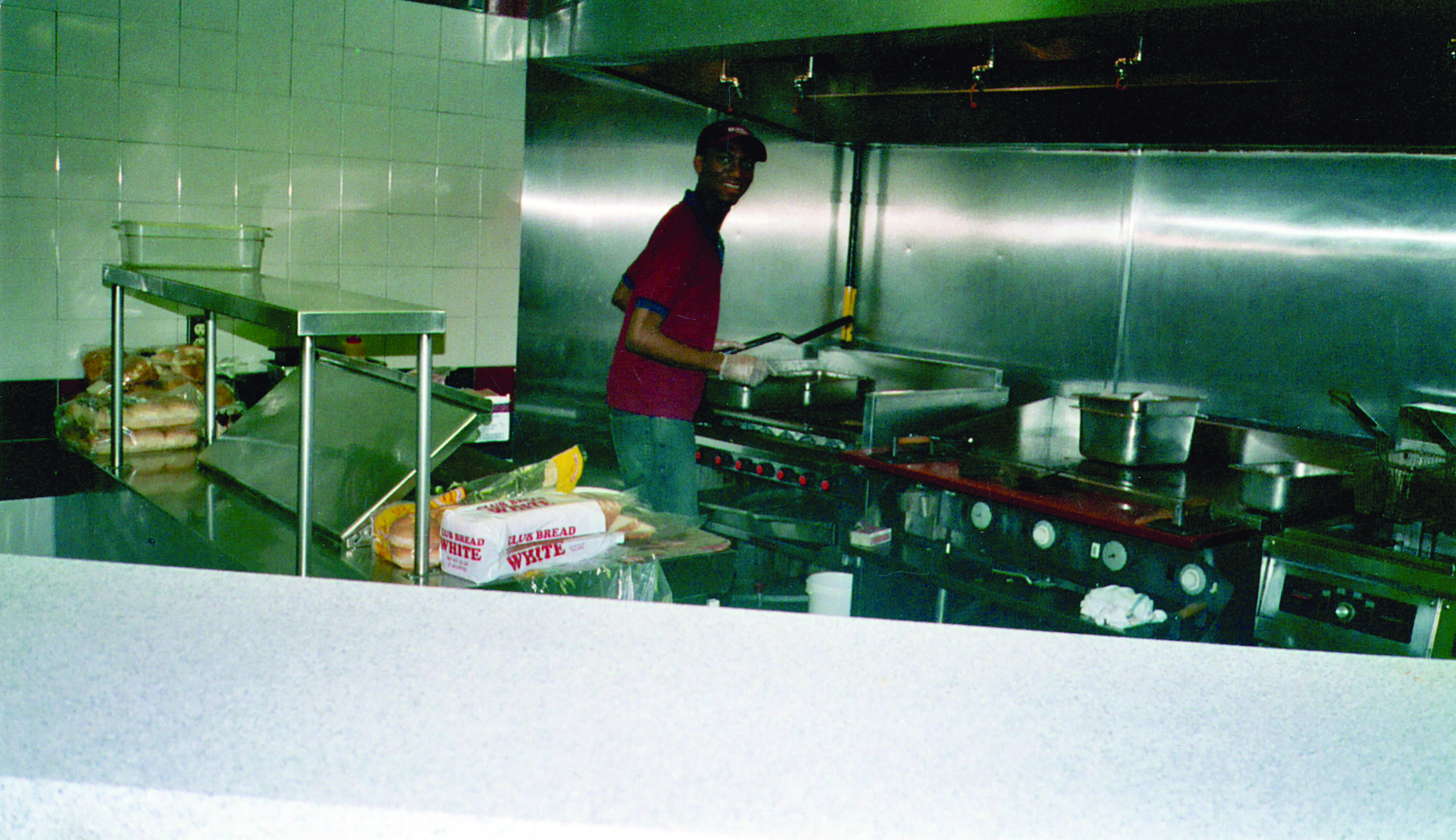 A man slightly turned, holding a spatula in front of a grill in a commercial kitchen