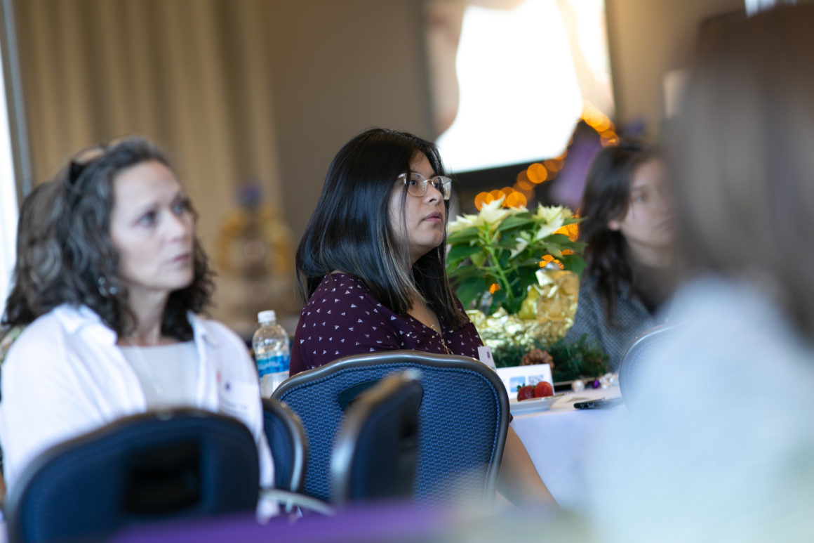 A woman sits at a table listening to a speaker give an address.