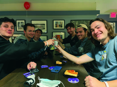 A group of six young men stand around a table and hold their drinks together for a toast
