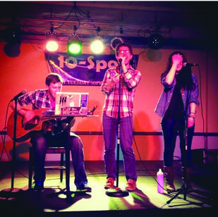 Three young men on a stage, two on the center and right hold a microphone, the one of the left sits and plays the guitar