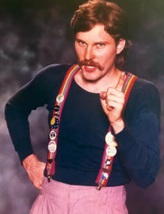Man with moustache and rainbow suspenders points at the camera