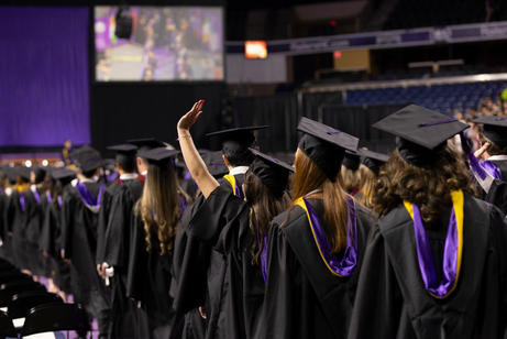 Student walking into a commencement ceremony wave to family seating in a large arena.