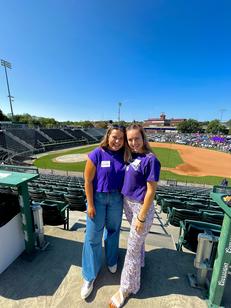 Two young woman posing and smiling with a baseball diamond behind them