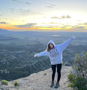 A young woman wearing a hooded Holy Cross sweatshirt stands atop a mountain overlooking a view of the valley