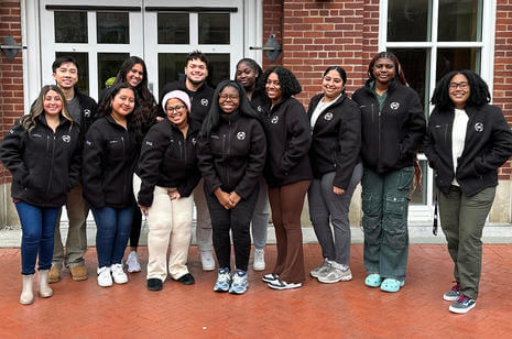 A group of students huddle together smiling outside a brick building