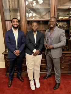 Three young men in formal attire pose with a bookshelf behind them