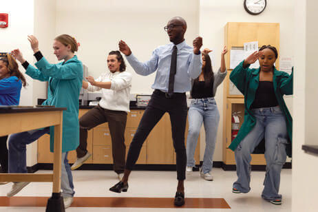 A man and four college students next to and behind him dance with their arms raised and right foot in the air.
