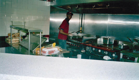 A man slightly turned, holding a spatula in front of a grill in a commercial kitchen