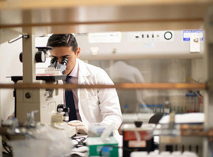 A student wears a white coat in a lab while looking through a microscope