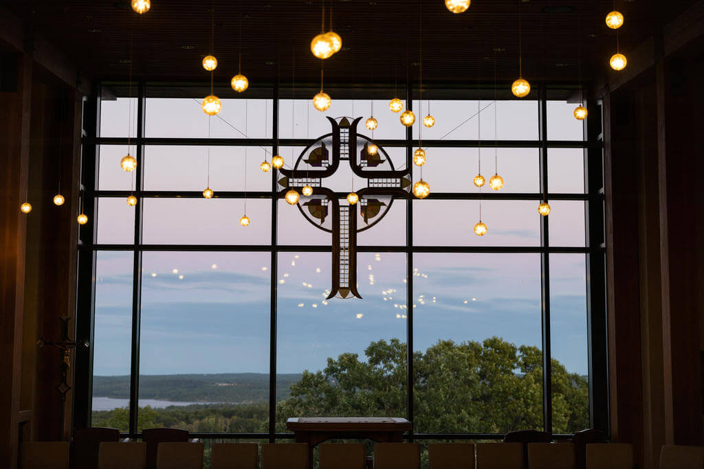 A cross hanging in front of a window looking out onto a forest and a lake