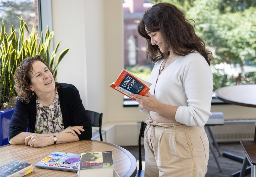 Seated women looks at women reading a book while standing