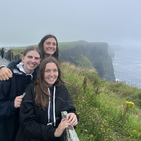 Three young women in matching sweatshirts stand along a cliff and the ocean in Ireland