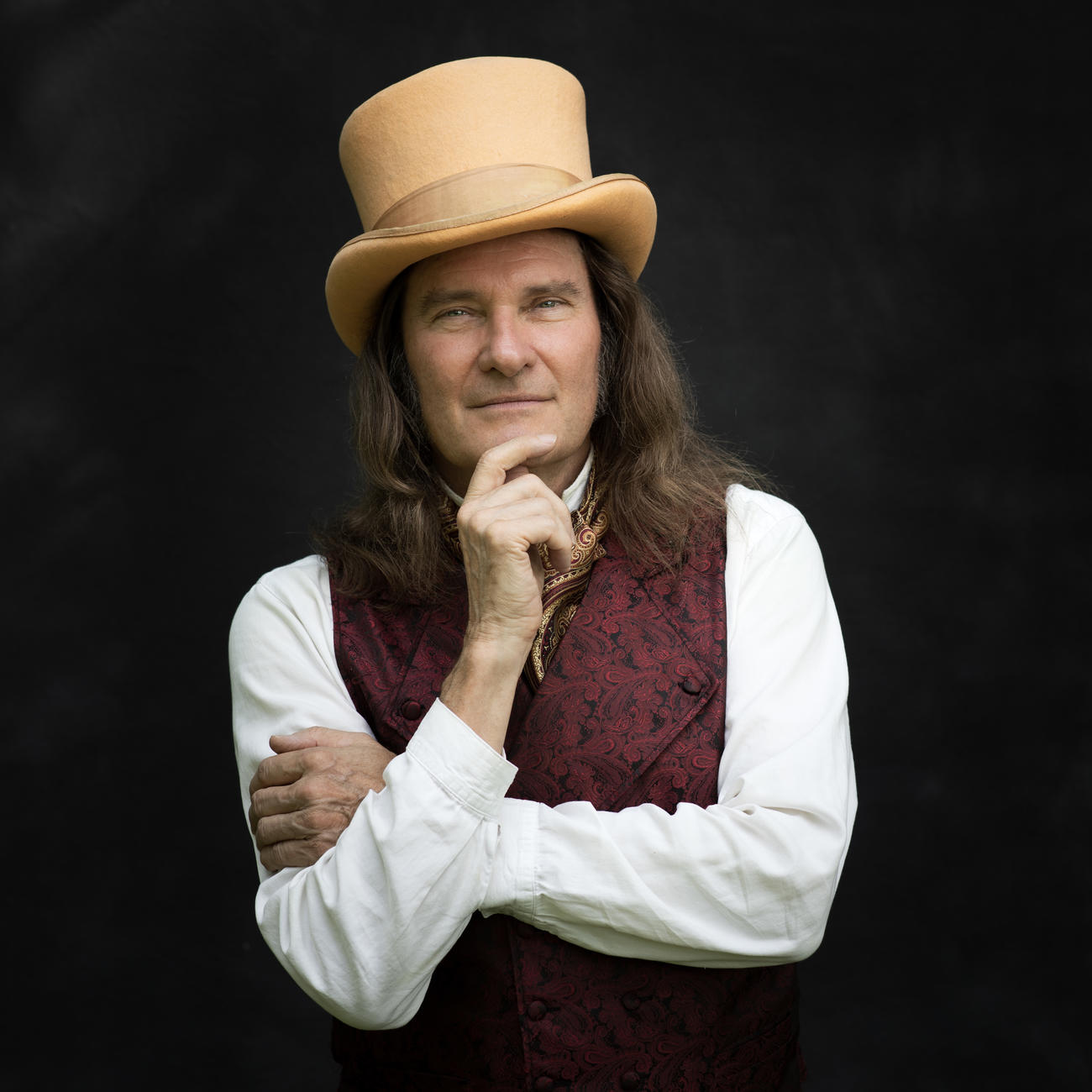 A white man in a top hat vest poses with his hand on his chin
