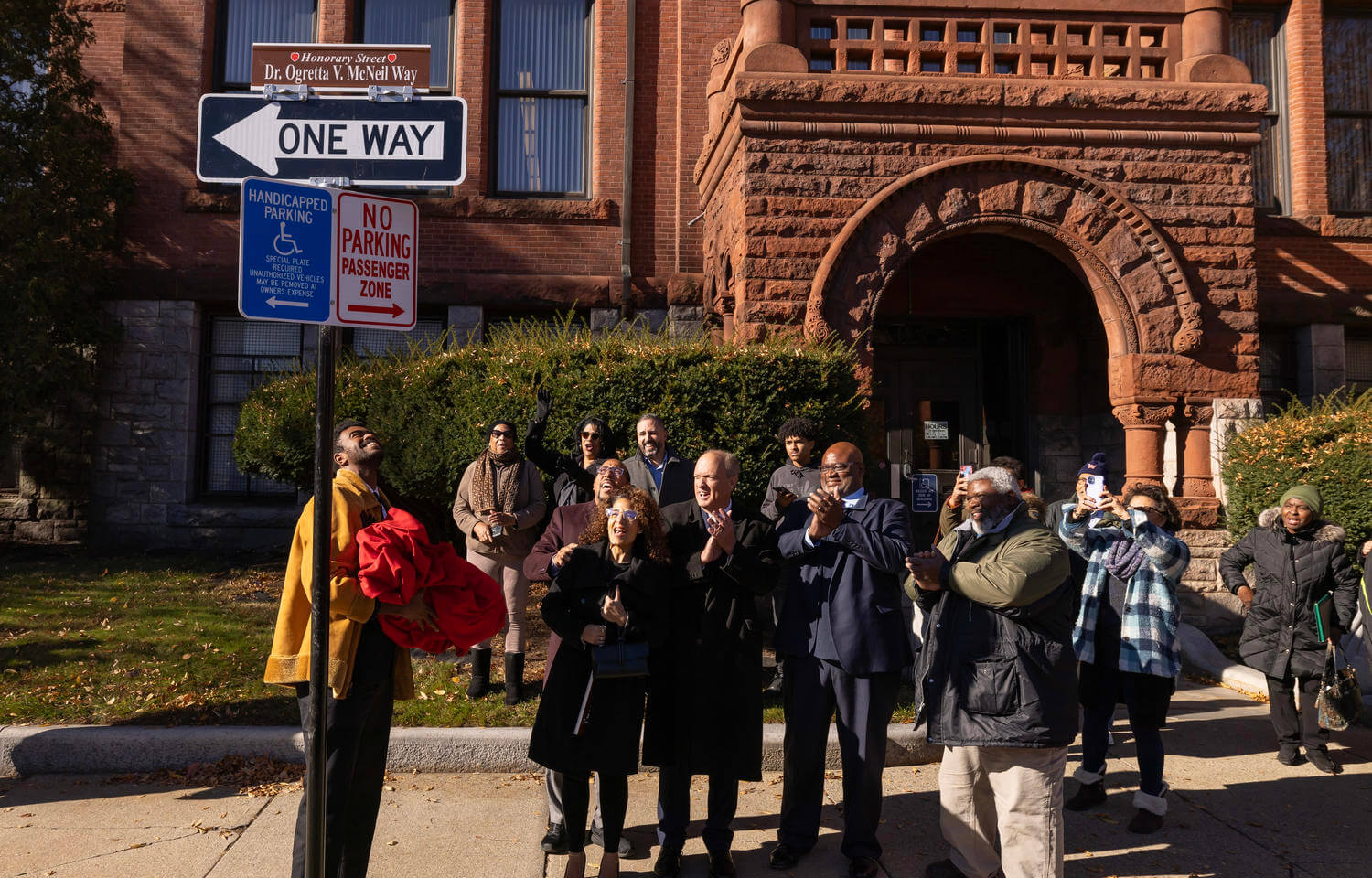 A group of people stand on the side of a street looking at a newly unveiled sign.