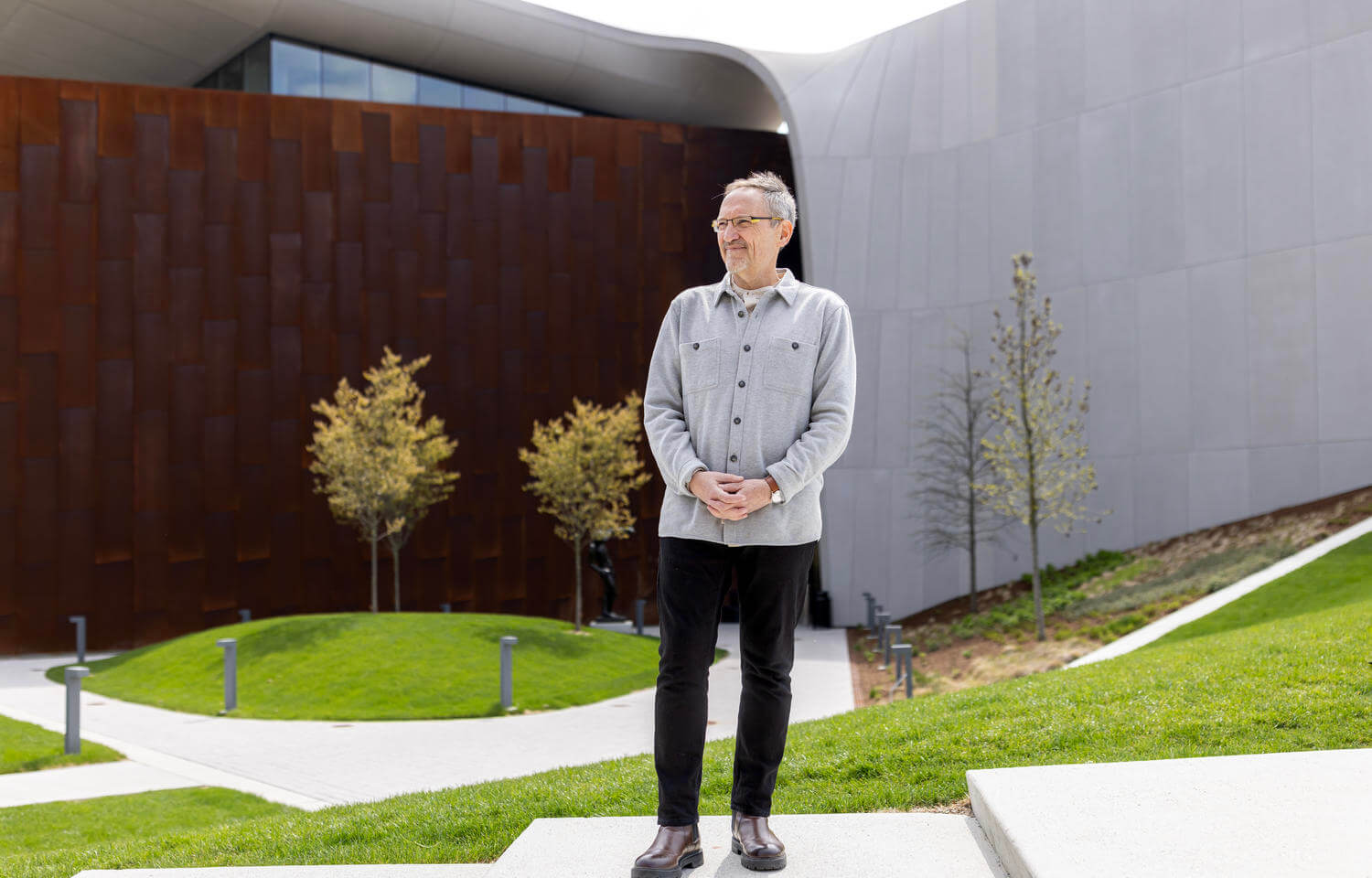 Man poses in front of building made of concrete and weathered steel walls.