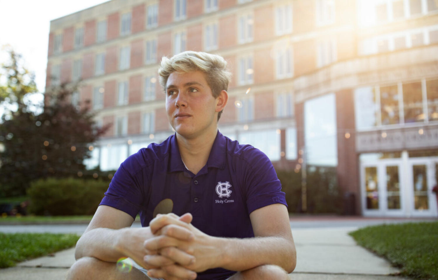 Man with blond hair sits with arms crossed in front of brick building with sun shining behind head.