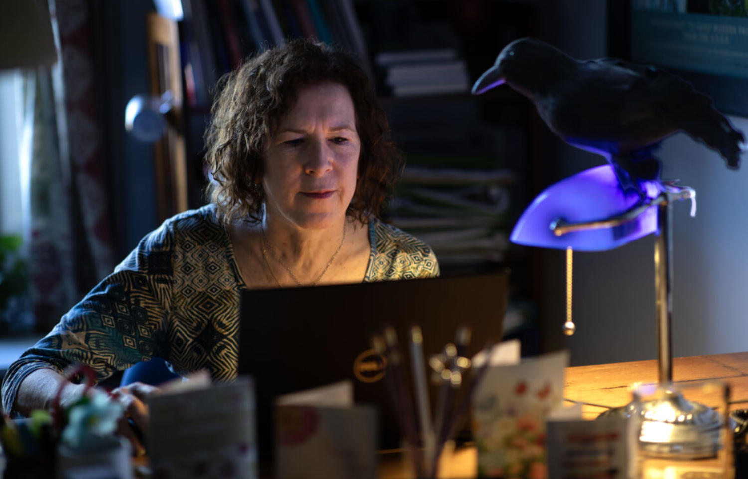 Woman sitting at a desk with a raven perched on top of a lamp.