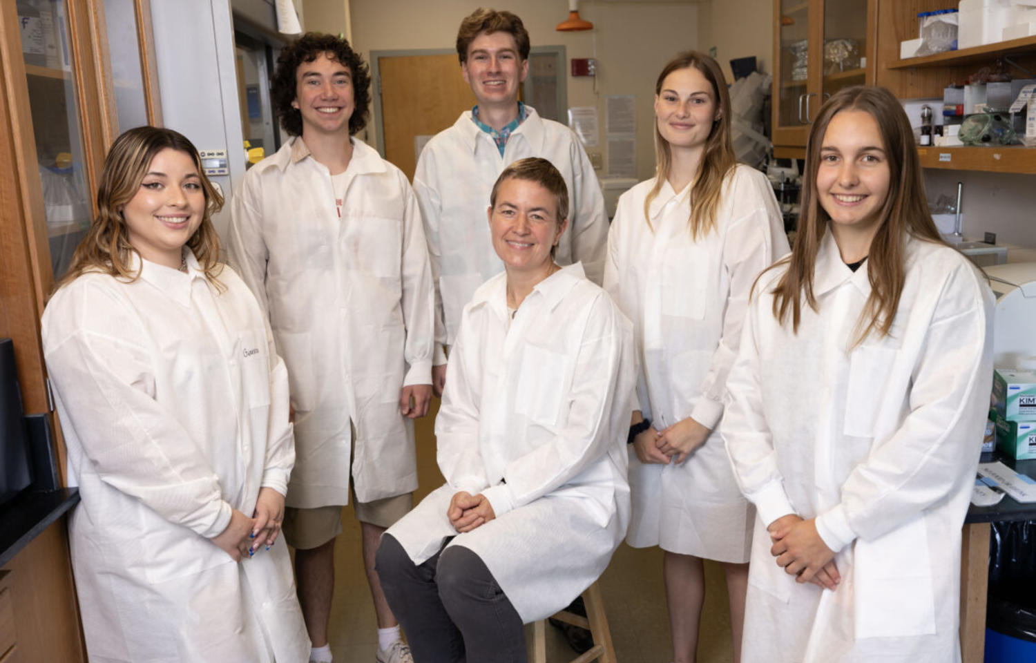 Biology students dressed in white lab coats stand behind their professor, who is seated in the center.
