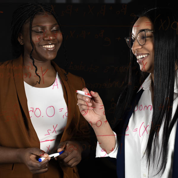 Two women write equations on a window. 