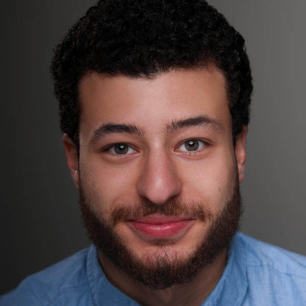 A portrait of a young man with a collared shirt and a beard staring with a slight grin