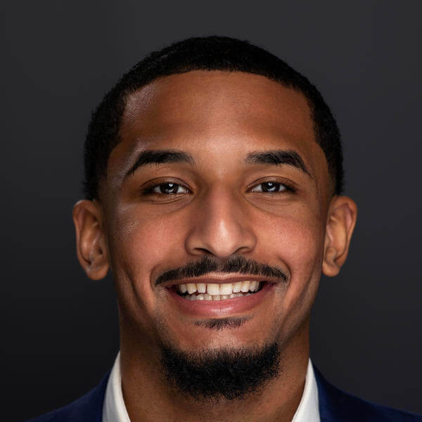 A portrait of a smiling young man with a goatee and short hair. He's wearing a white collared shirt and a dark jacket