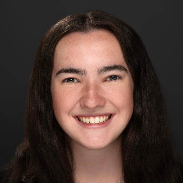 A portrait of a smiling young women with dark hair beyond her shoulders. She is earing a dark blouse.  