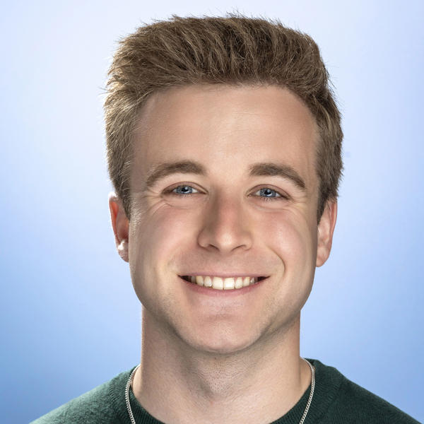 A smiling young man with wearing a green crew neck shirt and a necklace with a cross