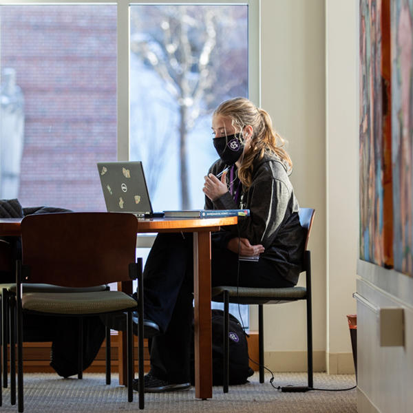 A student sits by a window on campus, working on their laptop.