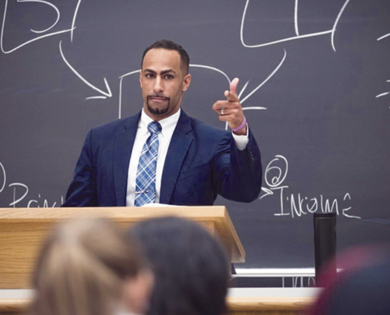 A man in a suit standing in front of a chalk board talking to students 