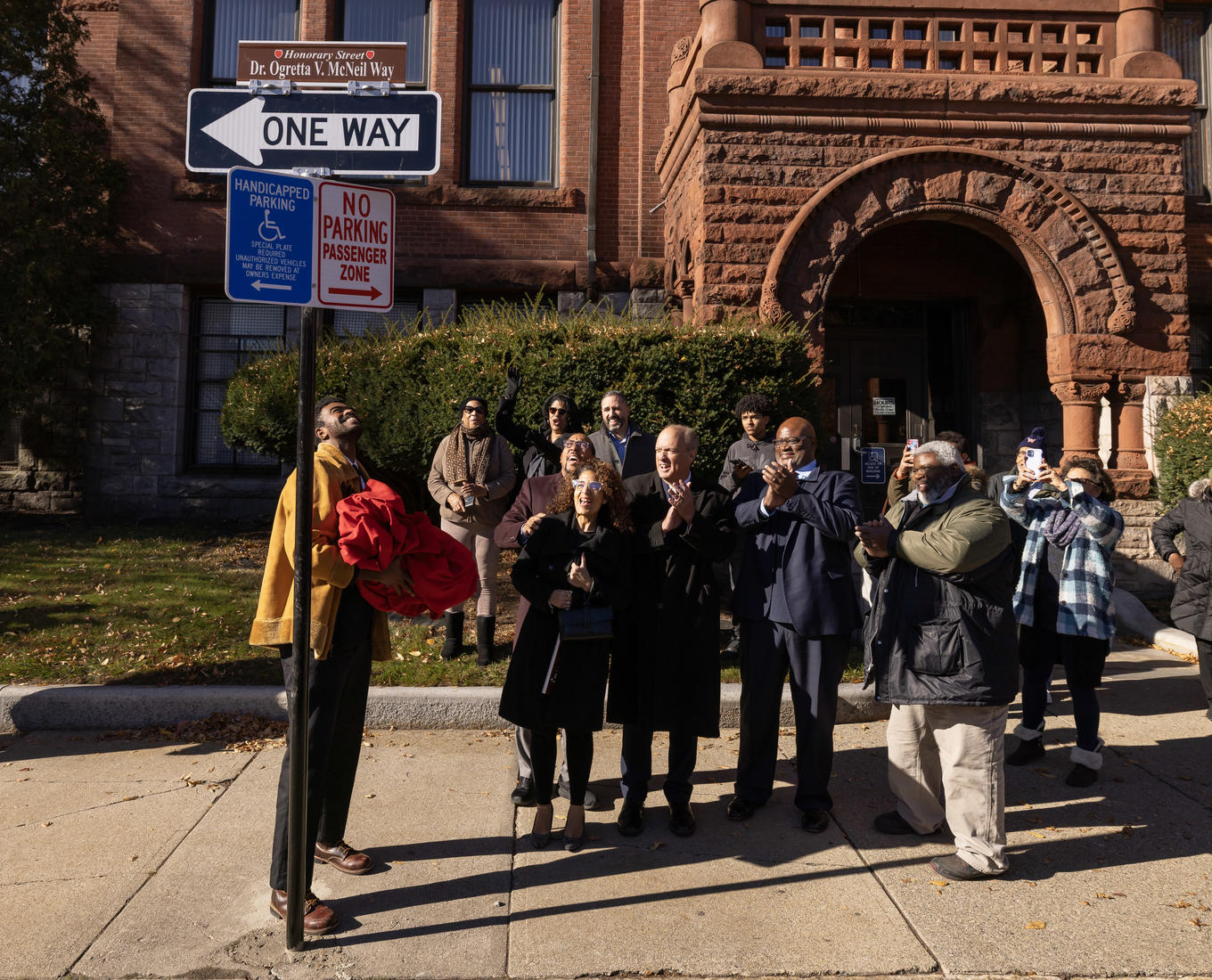 A group of people stand on the side of a street looking at a newly unveiled sign.