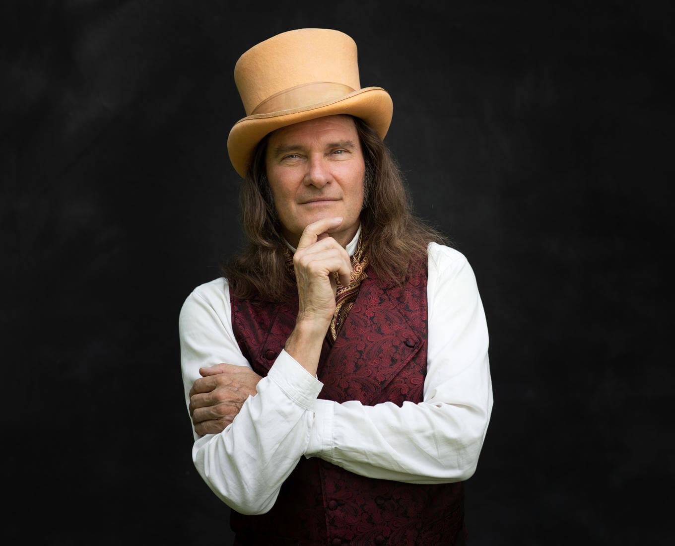 A white man in a top hat vest poses with his hand on his chin