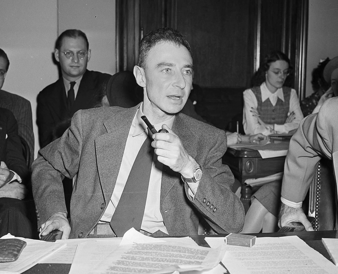 A man at a suit sits at a desk with paperwork covering it