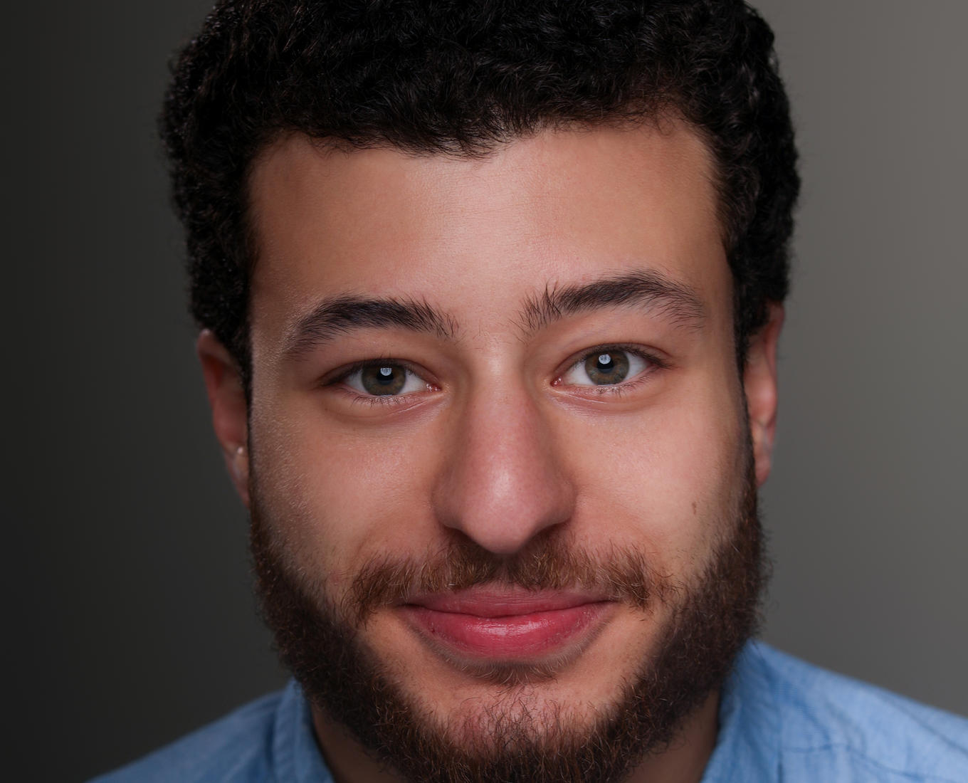 A portrait of a young man with a collared shirt and a beard staring with a slight grin