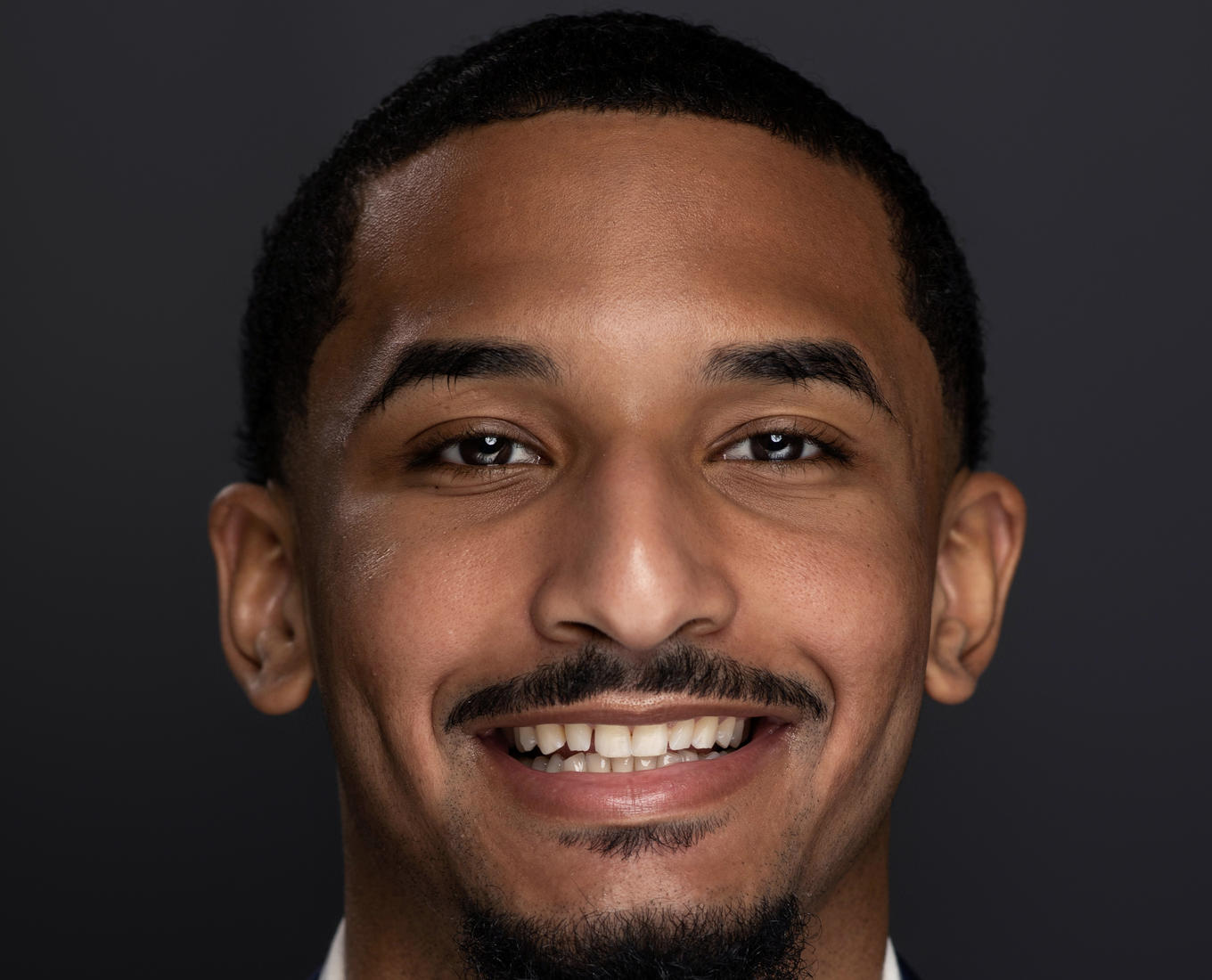 A portrait of a smiling young man with a goatee and short hair. He's wearing a white collared shirt and a dark jacket