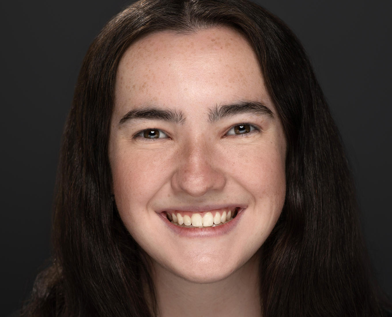 A portrait of a smiling young women with dark hair beyond her shoulders. She is earing a dark blouse.  