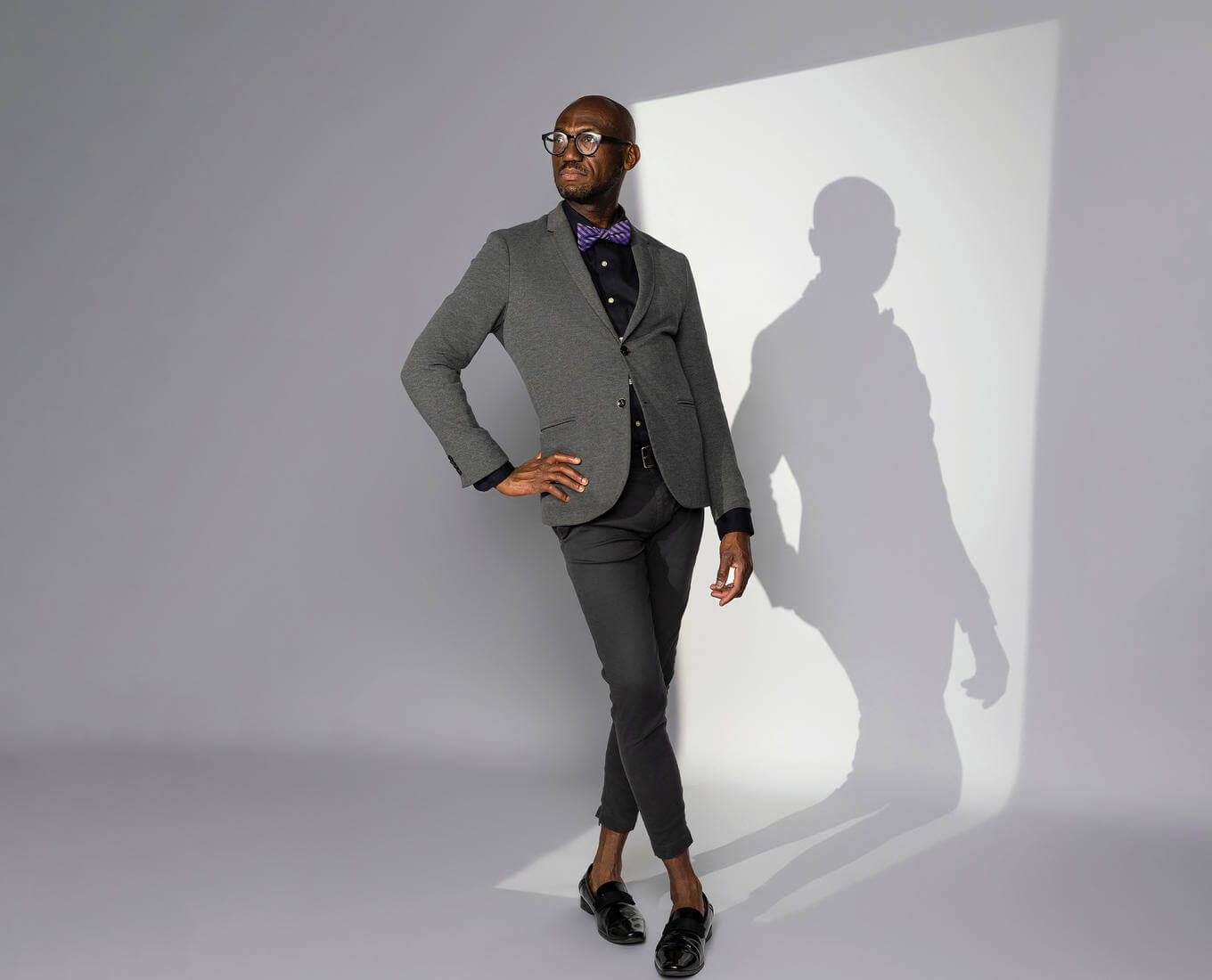 A man in a suit with a bow tie stands with his legs crossed and hand on hip, with his shadow in the background. 