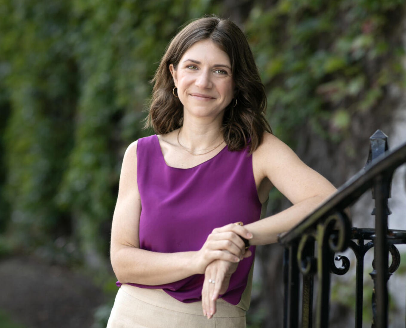 Woman dressed in purple shirt and tan skirt leans against a stair railing outside.