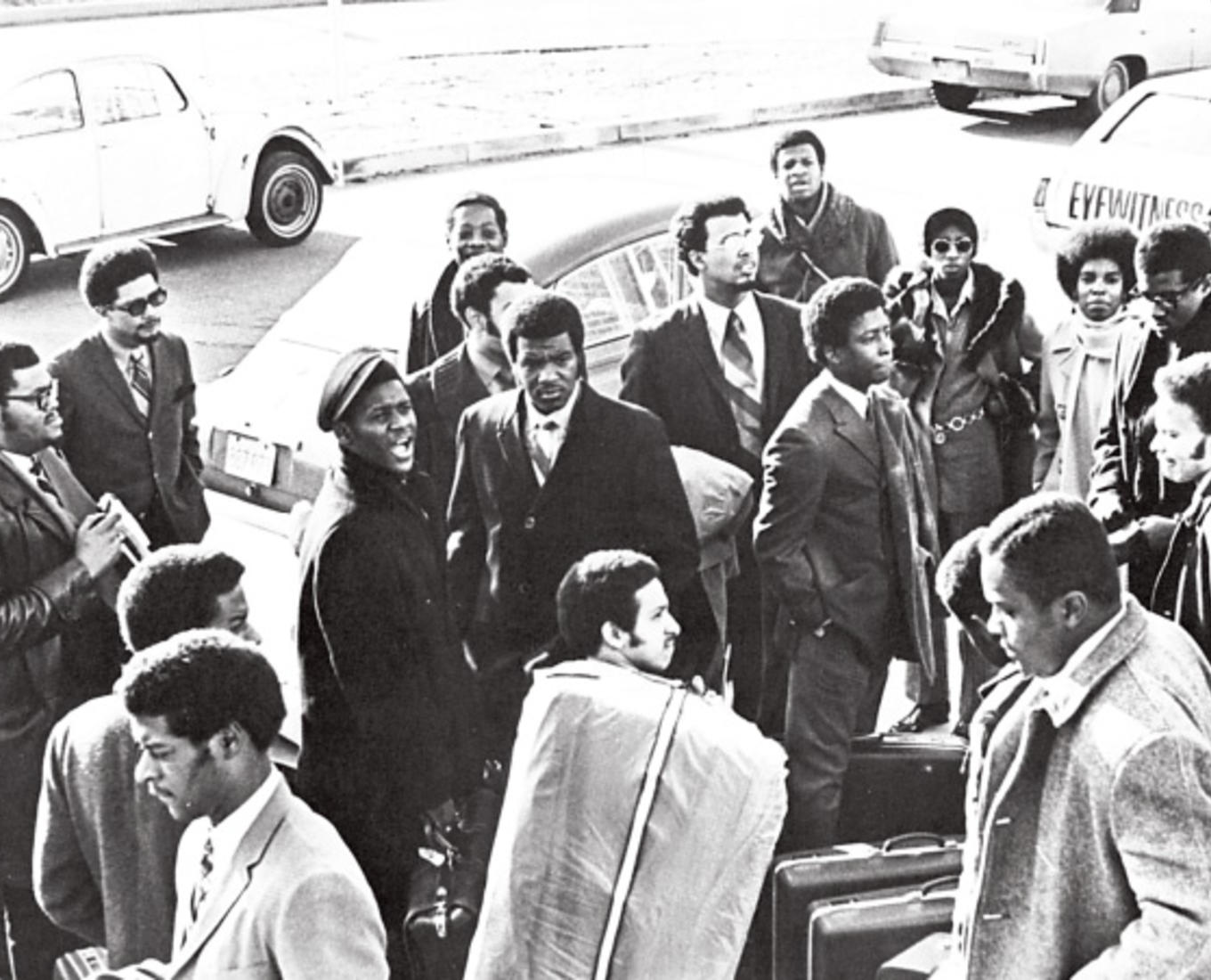 A group of mostly men gathered outside on a road by parked cars