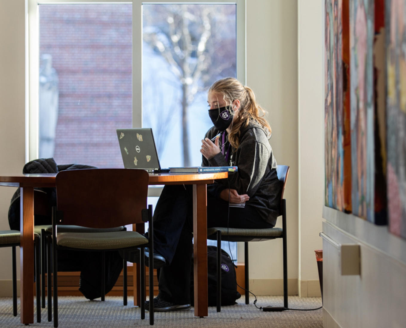 A student sits by a window on campus, working on their laptop.