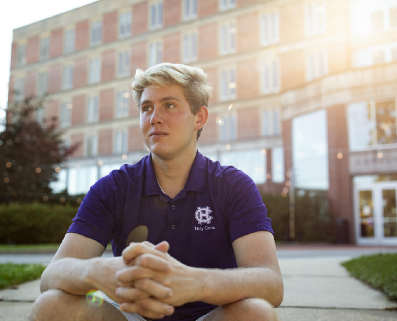 Man with blond hair sits with arms crossed in front of brick building with sun shining behind head.